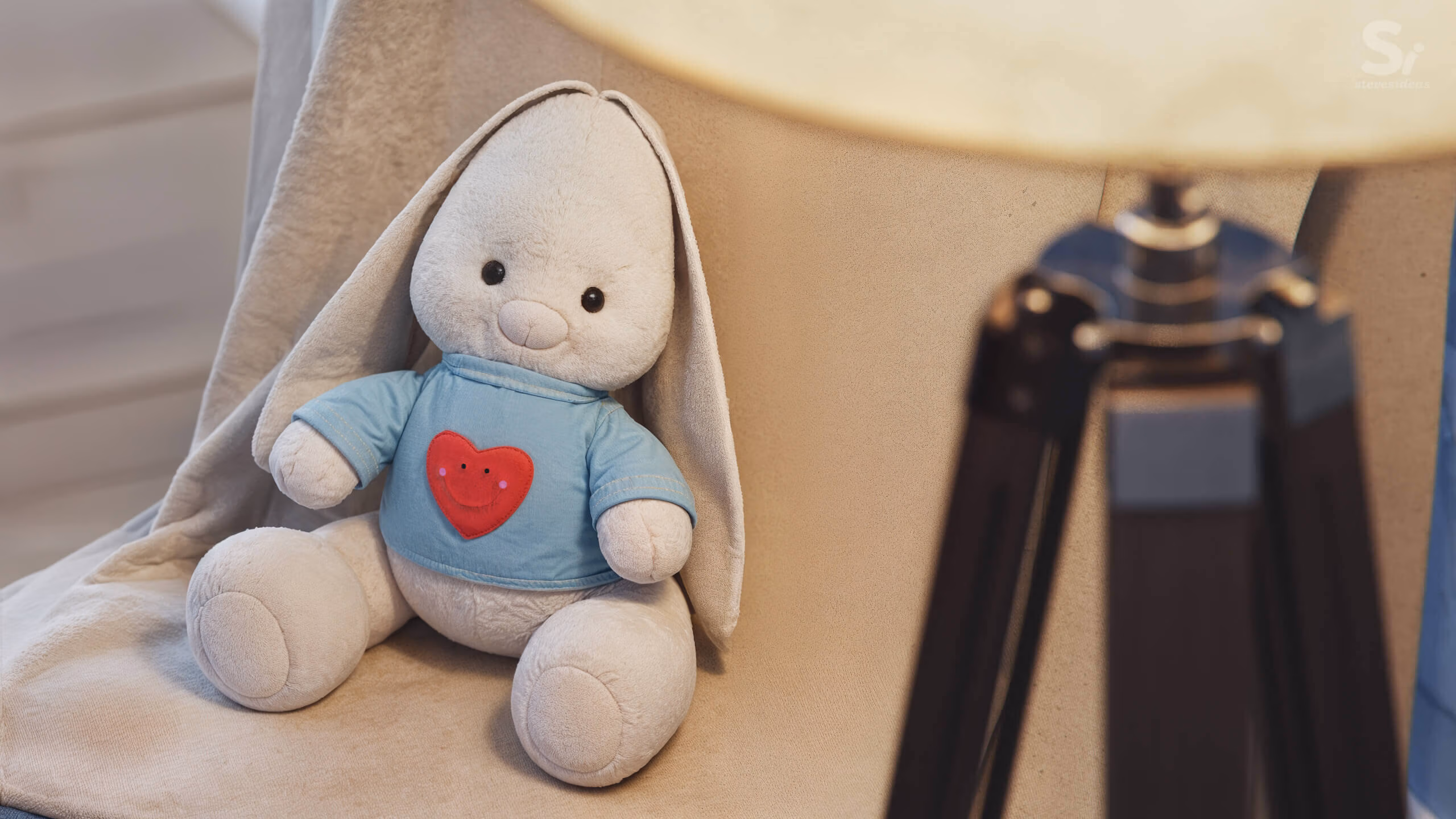 Close-up 3D render of a soft toy rabbit wearing a blue shirt with a red heart, sitting on a chair beside a warm floor lamp.